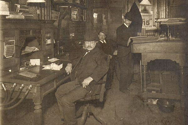 a man sitting wearing a suit and hat at a top heavy looking desk in 1930