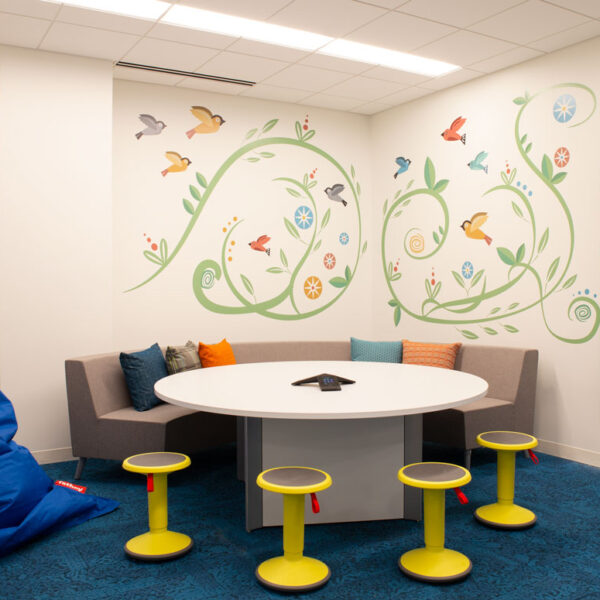 Playful meeting room with round table, yellow stools, and a wall mural of birds and vines, creating a cheerful family-friendly space at the YMCA