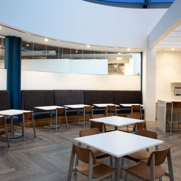Modern workplace café with gray upholstered booth seating, wood chairs, and white café tables designed for employee collaboration at the YMCA corporate office