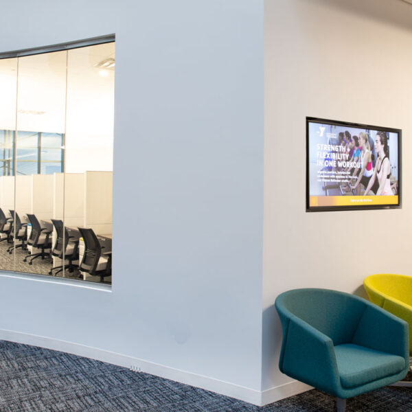 Office lobby seating area with blue and yellow lounge chairs, digital signage, and a view into a modern workspace at the YMCA headquarters